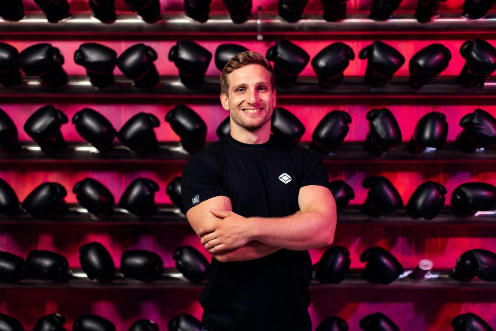 A smiling man in a gym stands confidently in front of a wall of boxing gloves.